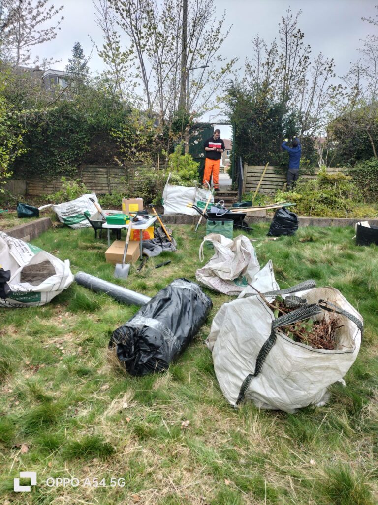 A full bag of pruned branches and anti-weed fabric lie on the grass. Tools, a garden volunteer, and piles of plants are in the background.