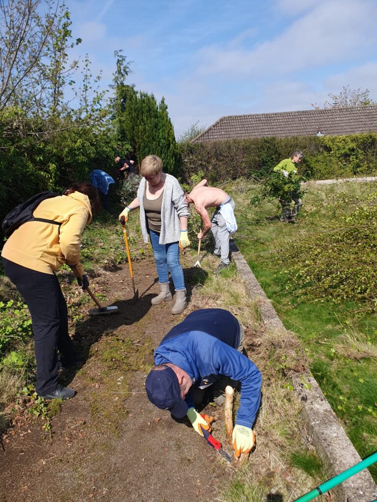 a group of people working to clear up a community garden site. There are people weeding, clearing paths and carrying cut branches.
