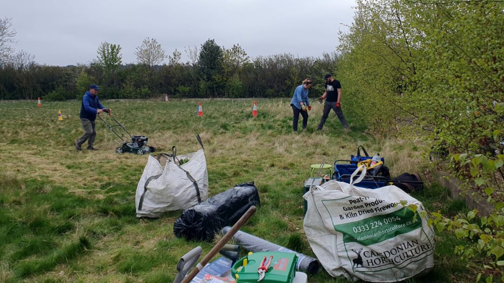 some large bags and tools lying on the grass. In the background, people are weeding and mowing grass.