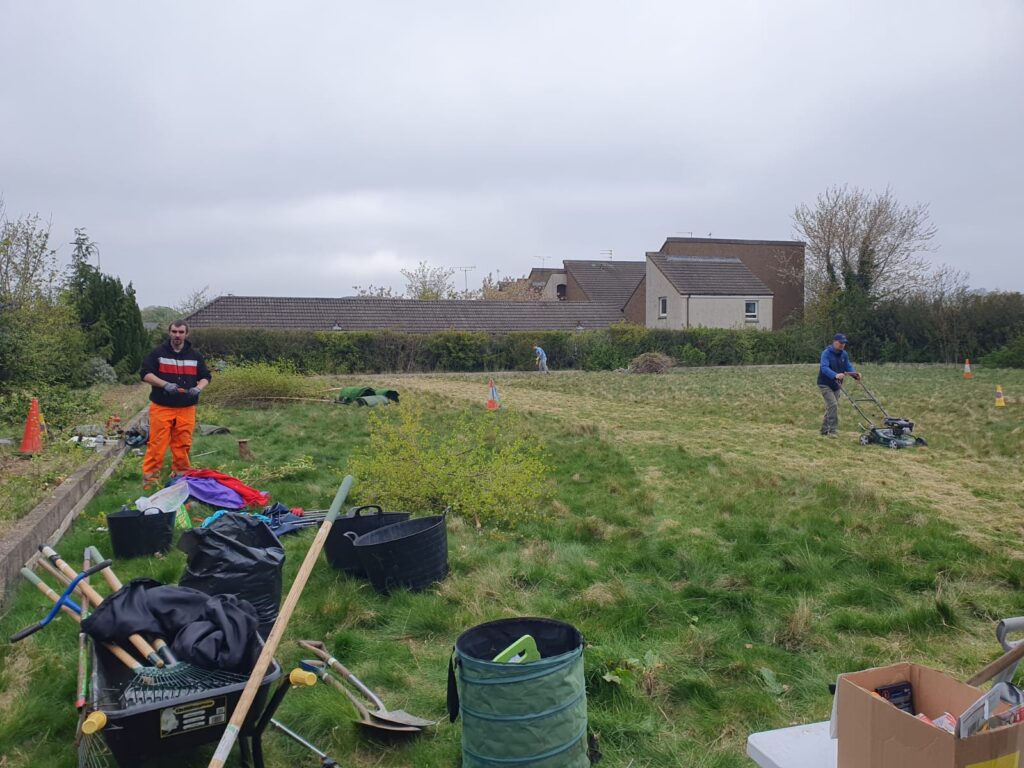 wide-angle view of an overgrown bowling green. On the right, a volunteer is mowing the grass. On the left, a volunteer stands behind tools and plant carriers.