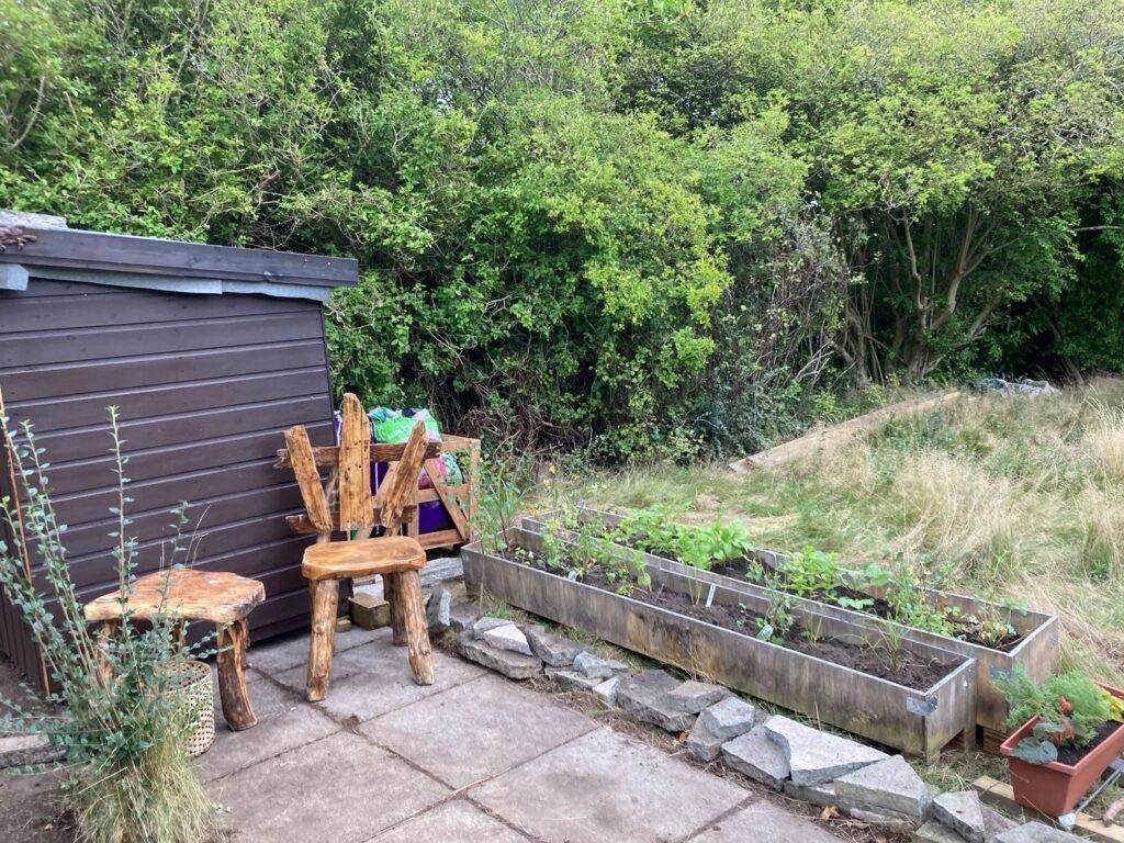 A small patio area with a handcrafted wooden chair and table in front of a small shed and two long planters