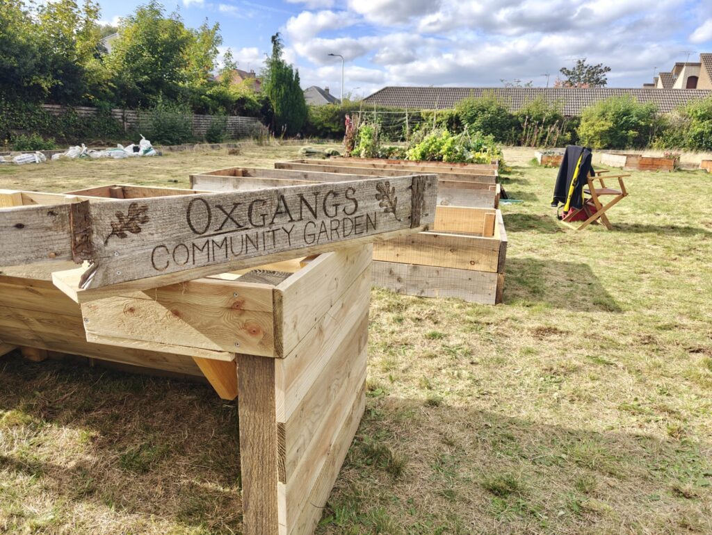 A sign saying "Oxgangs Community Garden" on a wooden mount, on top of a raised bed. The text is burnt into an old board, and there is an oak leaf on either side of the text.