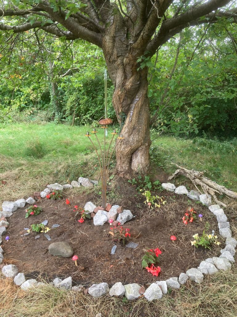 The twisted stem of a cherry tree behind a circle of light grey rocks containing perennial flowering plants, a candelabra and handcrafted wood and clay mushrooms.